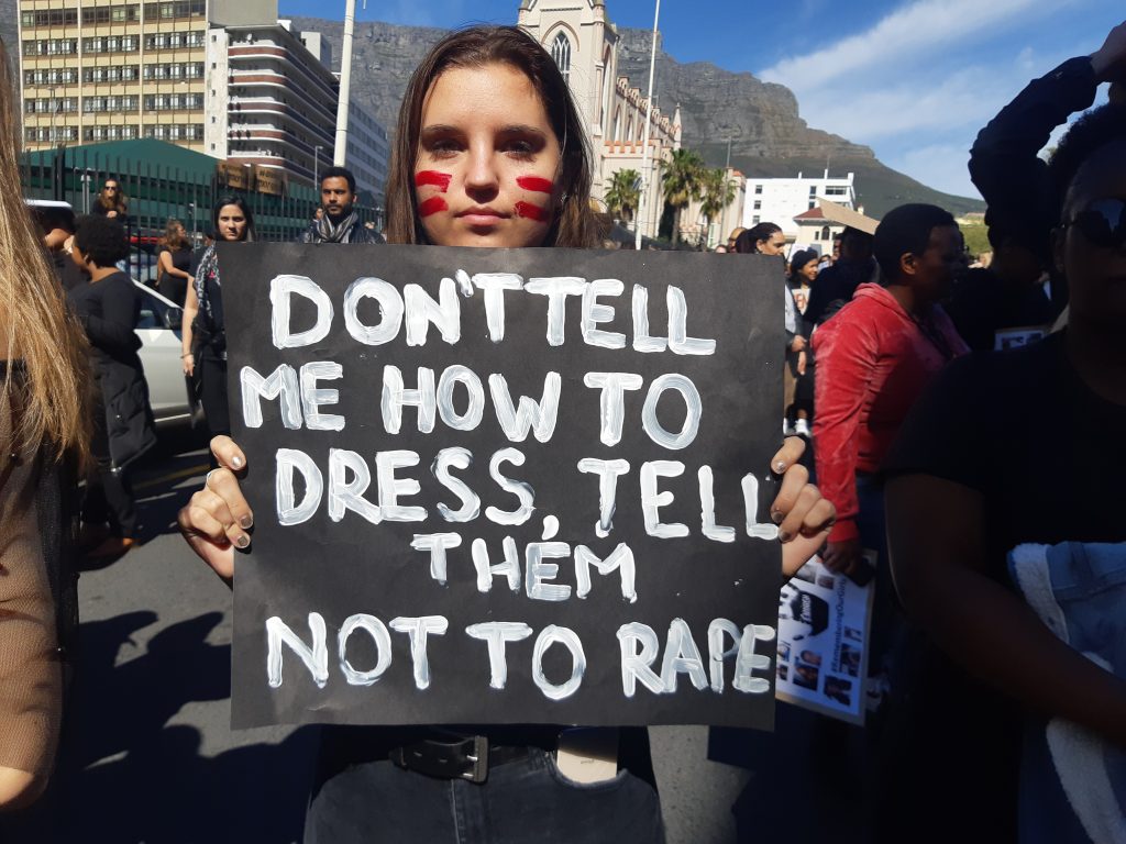 A young Woman holding a poster that reads "Don't tell me how to dress, tell them not to rape!".